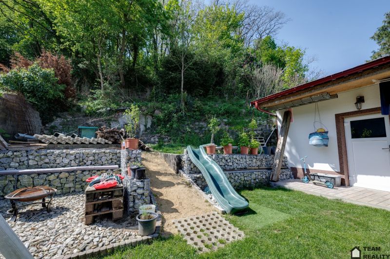 Rodinný dům v Potzneusiedlu s dvorkem, skluzavkou a kamennými zídkami obklopený zelení.