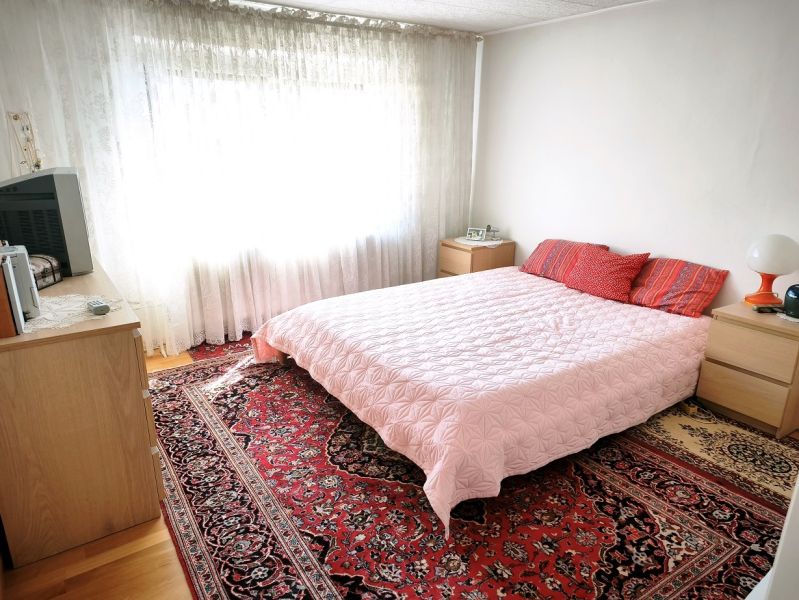 Ložnice v rodinném domě s postelí, červeným kobercem a světlými závěsy.