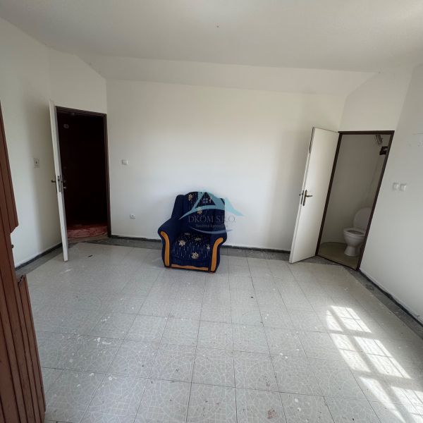 Pokoj s křeslem, bílými stěnami, dlažbou a dveřmi vedoucími na WC v chatě.