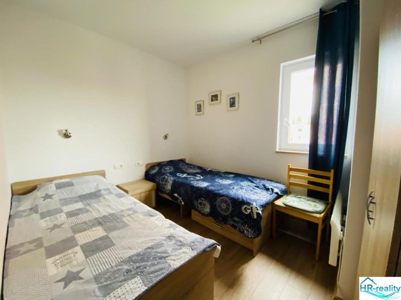 Dvě postele, židle, okno, obrazy na stěnách, podlaha s dřevěným dekorem.