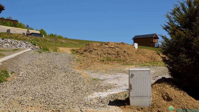 Rekreační pozemky v Nové Kelči, štěrková cesta, dřevěný dům, svahy a vegetace.