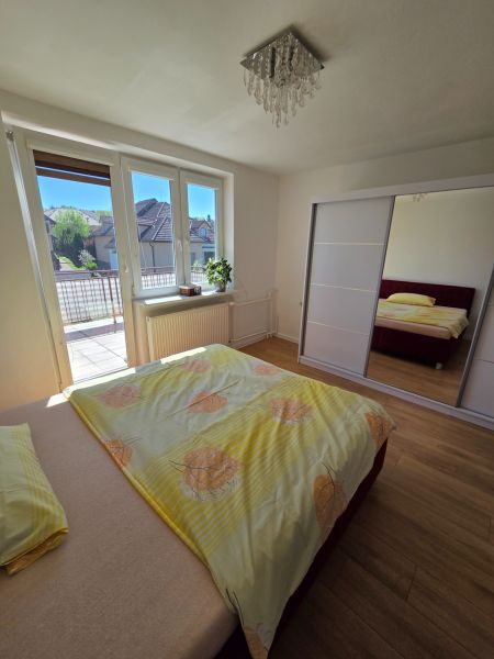 Ložnice v rodinném domě s postelí, skříní, oknem na balkon a podlahou s dřevěným dekorem.