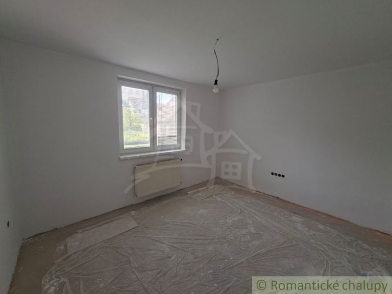 Prázdná místnost s plastovým oknem, radiátorem a odhaleným stropním svítidlem v rodinném domě.