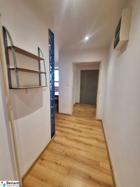 Chodba v 2-pokojovém bytě s dřevěnou podlahou a policí na stěně.