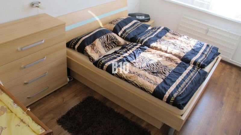 Ložnice se vzorovaným ložním prádlem, komodou a podlahou s dřevěným dekorem ve čtyřpokojovém bytě.