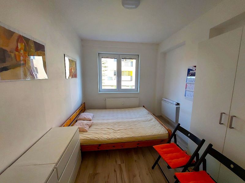 Garsonka s postelí, bílou skříní a podlahou s dřevěným dekorem, dvě sklápěcí židle.