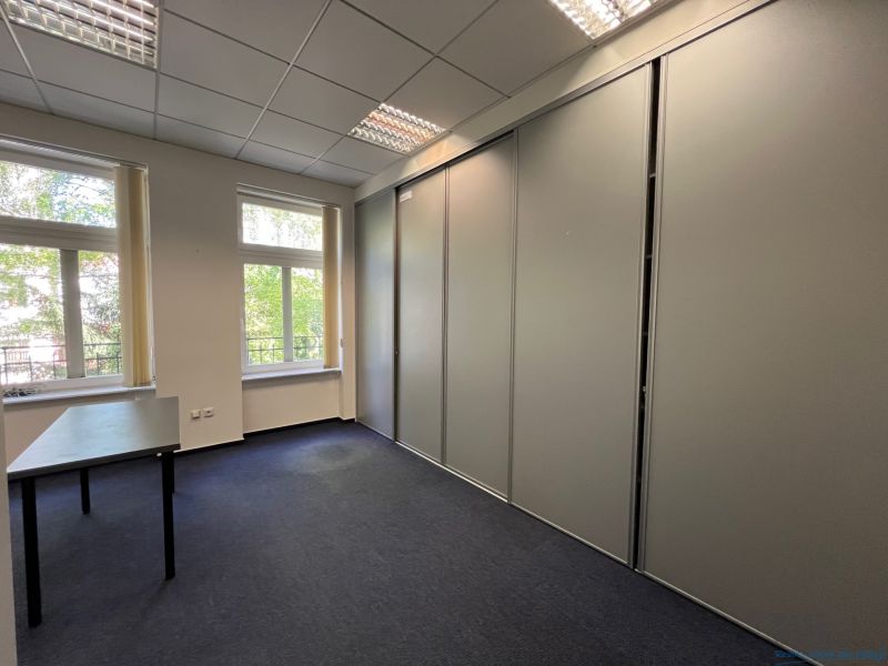 Kanceláře s velkým oknem a skříní, koberec, nábytek v neutrální barvě.