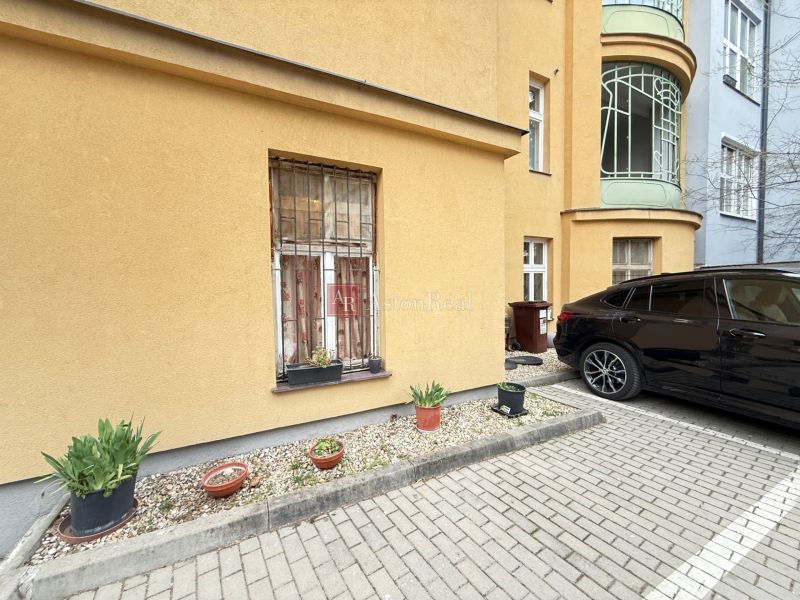 Parkování před budovou na Leškově v Bratislavě s květináči a zaparkovaným autem.