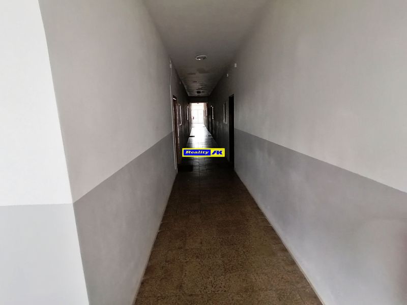 Dlouhá chodba s bílými stěnami a dlaždicovou podlahou v 1-pokojovém bytě.