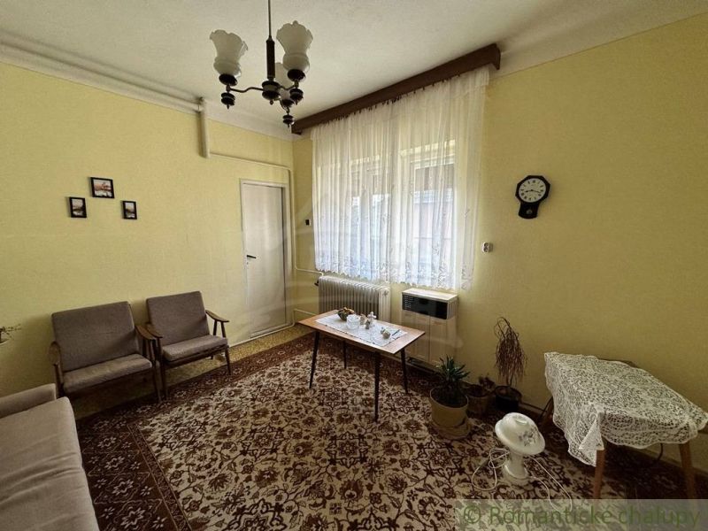 Obývací pokoj v rodinném domě s kobercem a záclonami. Retro nábytek doplňuje útulnou atmosféru.