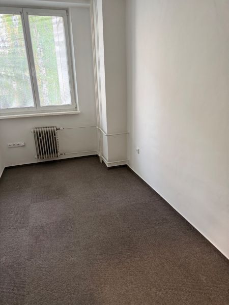 Kancelář s kobercem, radiátorem a oknem se žaluziemi.