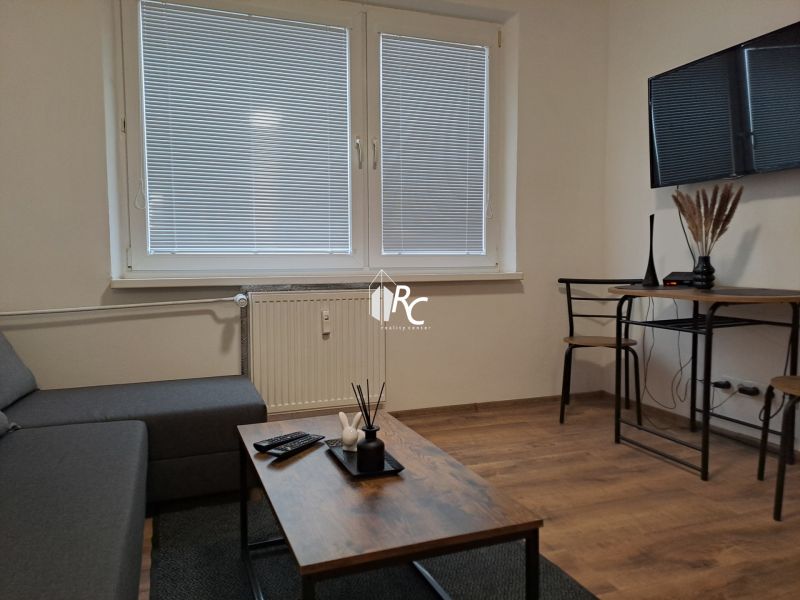Obývací pokoj v jednopokojovém bytě, šedo-hnědá pohovka, stolek a podlaha s dřevěným dekorem.