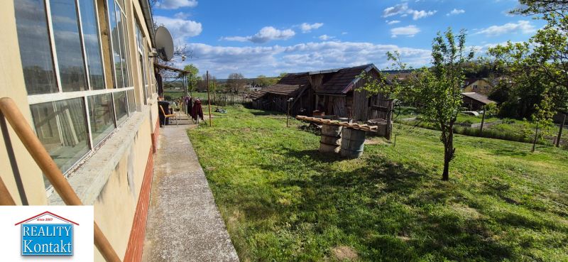 Rodinný dům v Dedince s velkou zahradou, zelení a starší budovou v pozadí.
