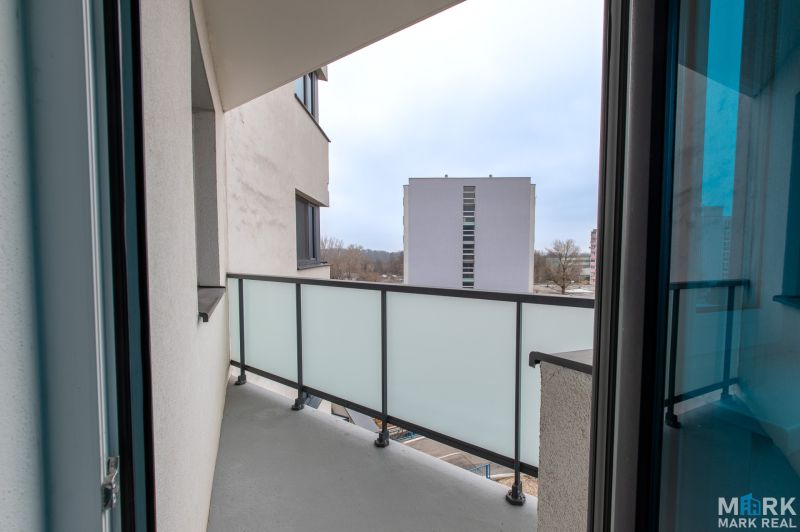 Balkon v 2-pokojovém bytě s výhledem na okolní budovy přes skleněné zábradlí.