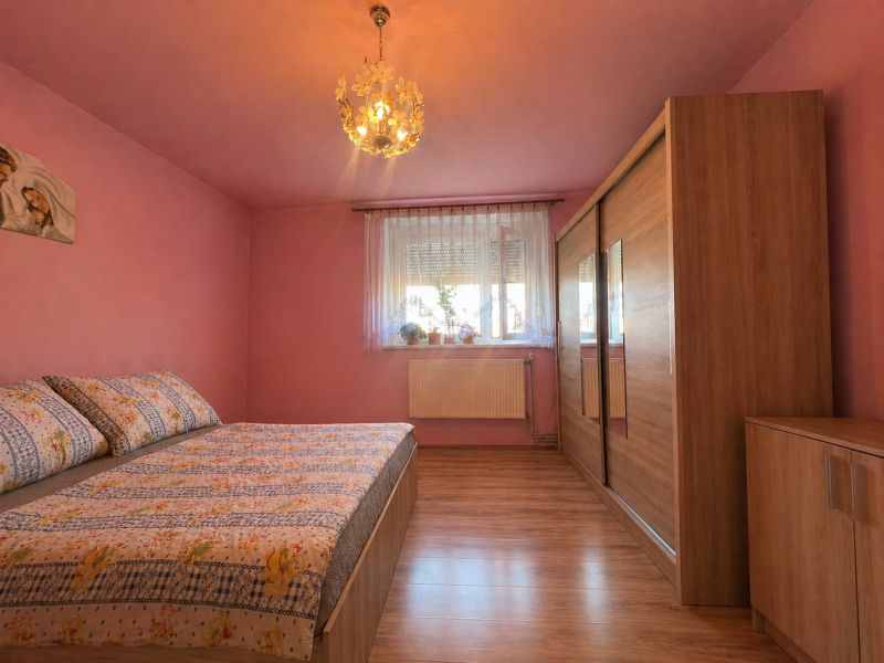 Ložnice s růžovými stěnami, postelí a šatníkem, podlaha s dřevěným dekorem.