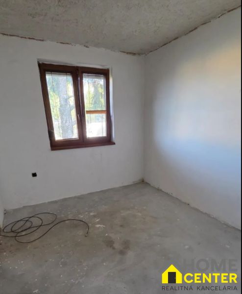 Nedokončený pokoj s oknem v 3-pokojovém bytě, světlá stěna a betonová podlaha.