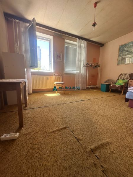 Pokoj v 2-pokojovém bytě s kobercem, otevřeným oknem a jednoduchým nábytkem.
