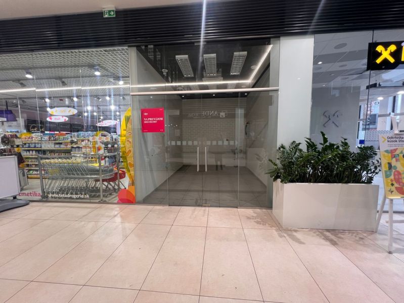 Obchodní prostory s prosklením a rostlinami, vedle drogerie v nákupním centru.