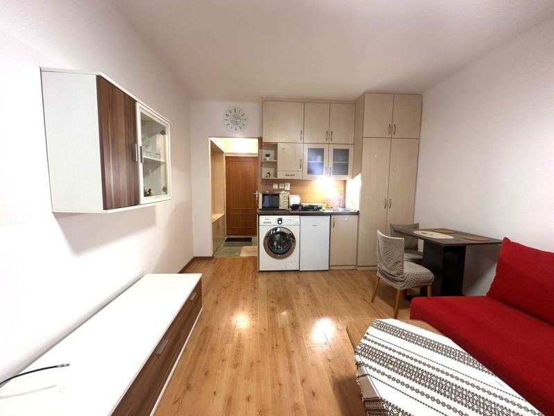 Rekreační apartmán s vestavěnou kuchyní, červenou sedačkou a podlahou s dřevěným dekorem.
