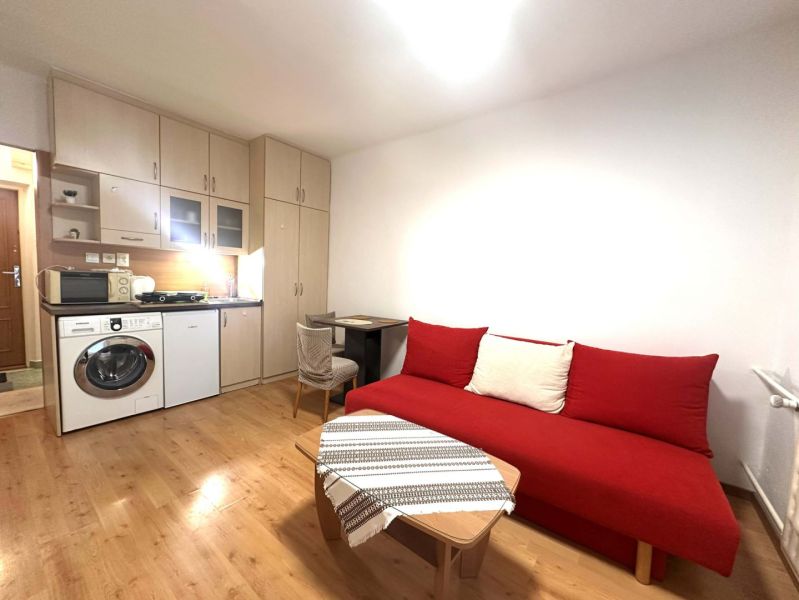 Rekreační apartmán s červenou sedačkou, kuchyňským koutem a podlahou s dřevěným dekorem.