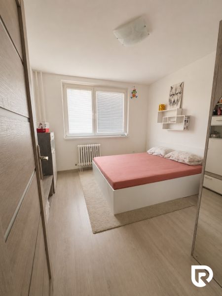 Ložnice ve 3pokojovém bytě s podlahou s dřevěným dekorem a jednoduchou postelí.