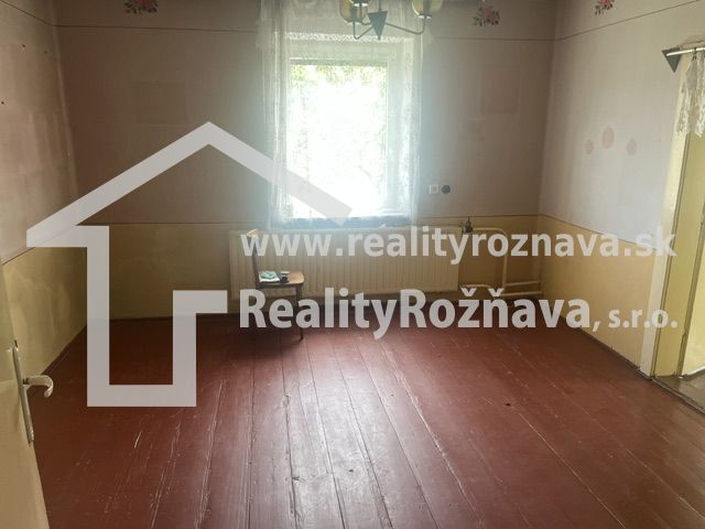 Pokoj v rodinném domě s dřevěnou podlahou, stěny pastelových barev, okno s bílou záclonou.