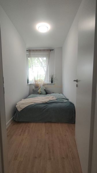 Úzká ložnice s postelí a oknem, podlaha s dřevěným dekorem, světlý interiér.