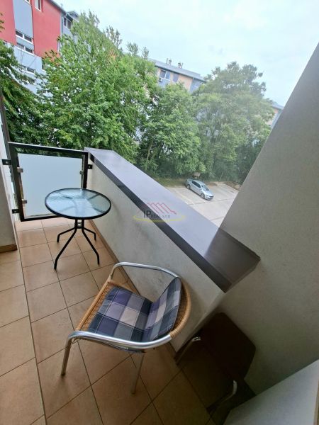 Balkon garsonky s výhledem na zeleň a parkoviště, místnost se stolem a židlí.
