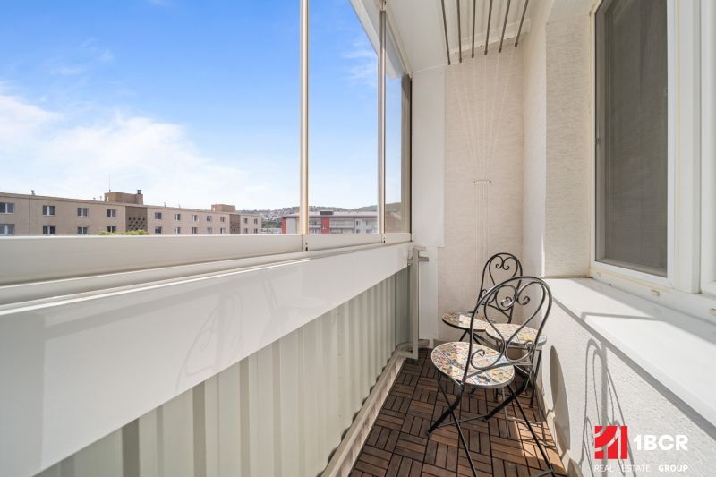 Zasklený balkon s dřevěným dekorem podlahy v 3-pokojovém bytě se stolem a židlemi.