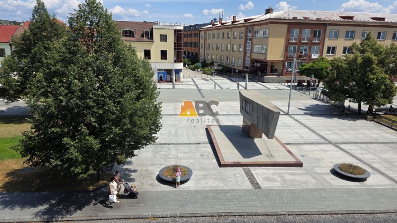 Náměstí SNP ve Zvolenu s monumentem a chodci, obklopené kancelářemi a stromy.
