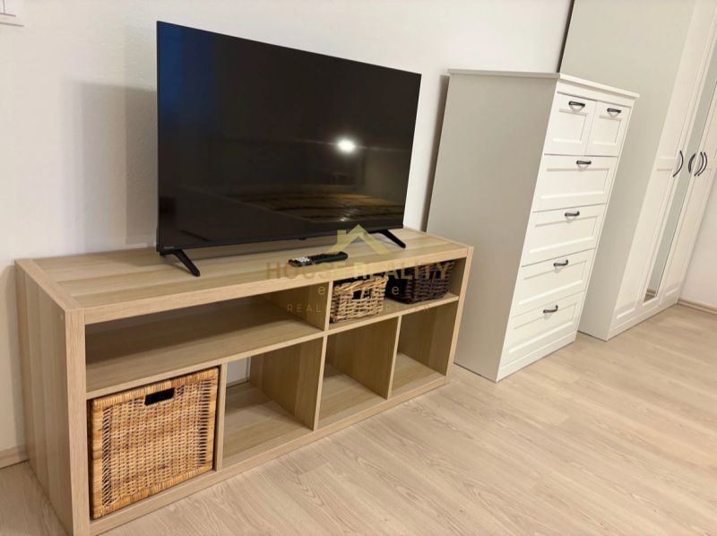Televize na poličce s košíky, vedle komody a skříně, podlaha s dřevěným dekorem.