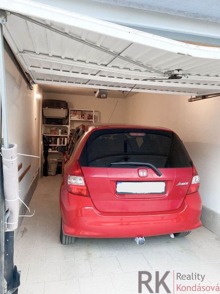 Červené auto zaparkované v garáži s regály na stěně, podlaha z dlažby.