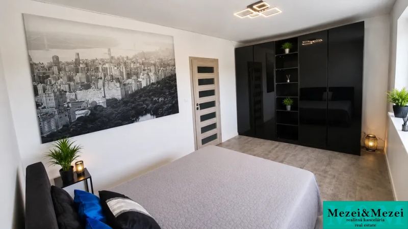 Ložnice s černou šatní skříní, velkou fotografií a pokojovými rostlinami.