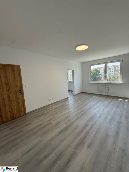 Pokoj v třípokojovém bytě s podlahou s dřevěným dekorem, bílými stěnami a oknem.