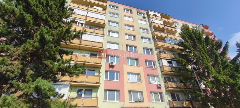 Bytový dům v Lučenci s balkóny, lemovaný zelenými stromy.