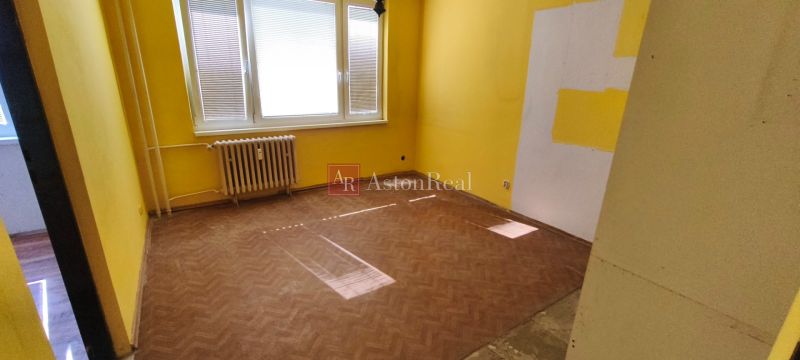 Pokoj s podlahou s dřevěným dekorem, žluté stěny, radiátor, jedno okno.