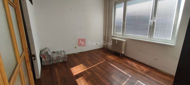 Pokoj s křeslem, okny a podlahou s dřevěným dekorem v 1+kk bytě.