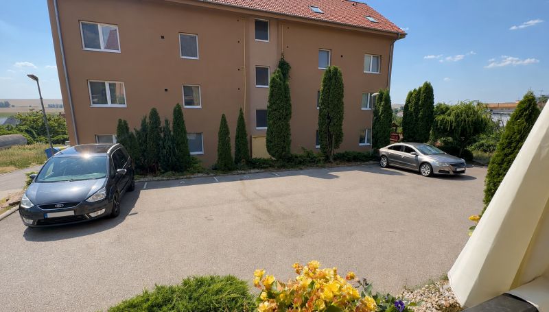 Parkoviště před bytovkou ve Velkém Lapáši s auty a zelení v okolí.