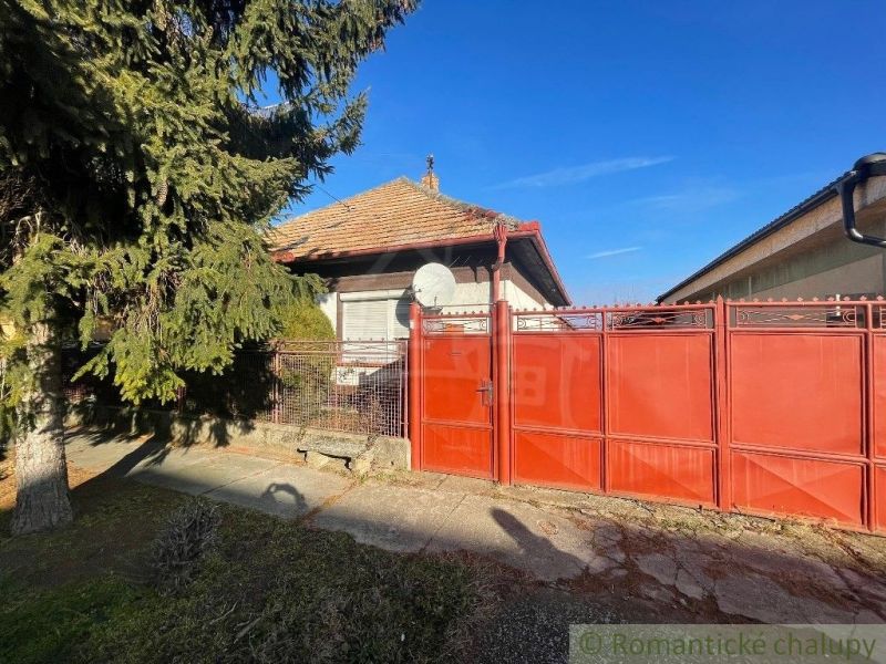 Červená brána a plot s rodinným domem v Tvrdošovcích, obklopený zelení.