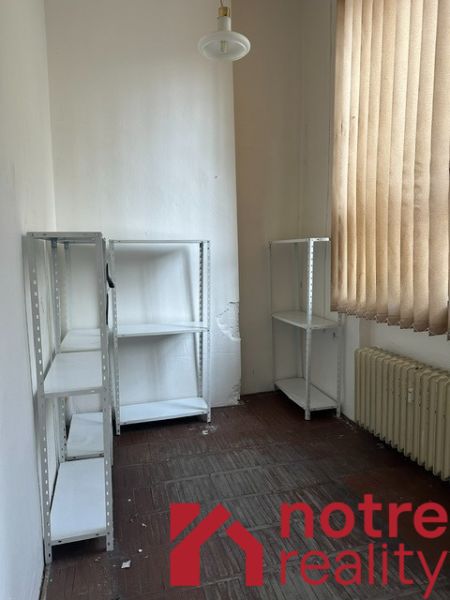 Kancelář s regály, radiátorem a závěsy, podlaha s dřevěným dekorem.