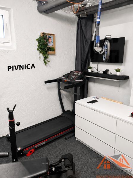 Fitness koutek s běžeckým pásem, televizorem a činkovou lavicí ve sklepě 3-pokojového bytu.