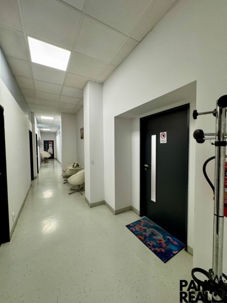 Chodba obchodních prostor se světlými stěnami, dekorativními rohožkami a nástěnnými světly.