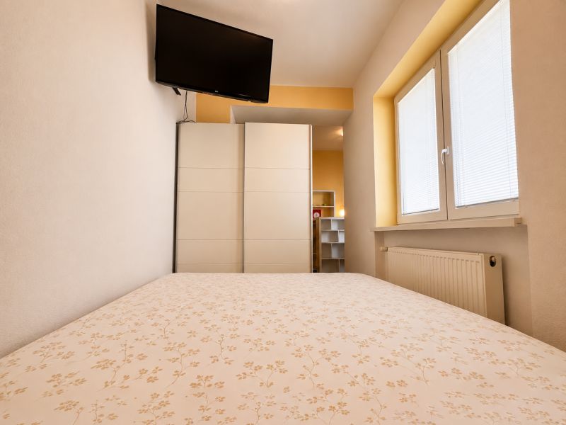 Ložnice v garsonce s televizí, skříní a oknem s roletami.