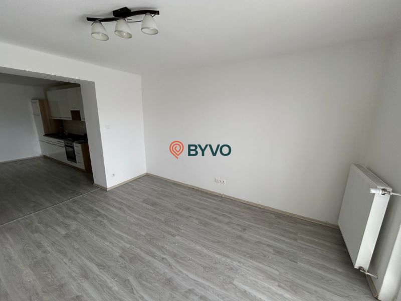 Obývací pokoj třípokojového bytu s podlahou s dřevěným dekorem a kuchyní na levé straně.