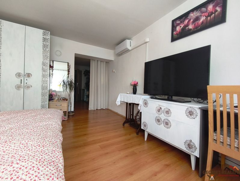 Ložnice s podlahou s dřevěným dekorem, televizorem, skříňkou a květinovou dekorací.