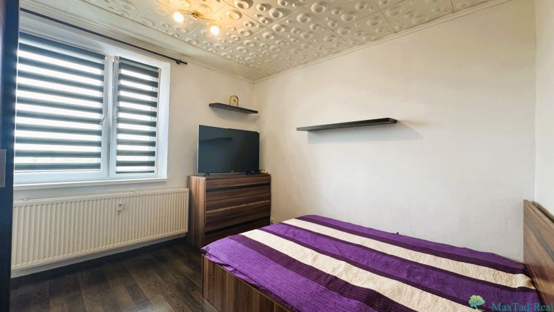 Ložnice ve 4pokojovém bytě s postelí, televizí, podlahou s dřevěným dekorem a moderními žaluziemi.