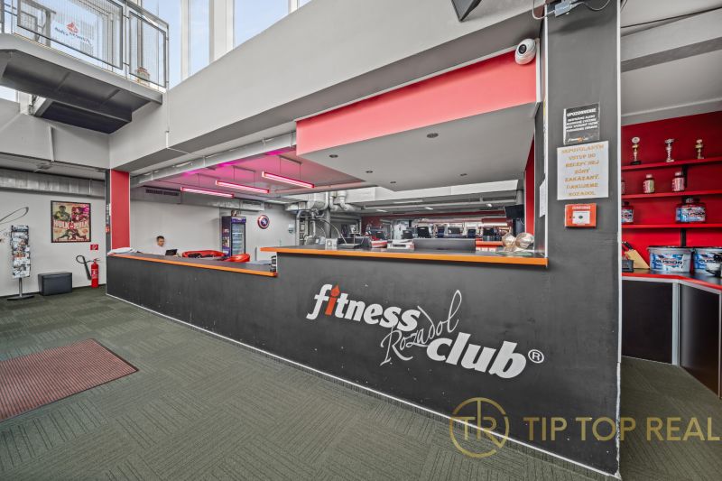 Recepce fitness klubu s nápisem, červeným osvětlením a dekorativními trofejemi na zemi zeleného odstínu.