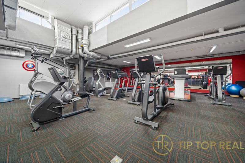 Fitness centrum s posilovacím vybavením, zrcadly a vzduchotechnikou v obchodních prostorech.