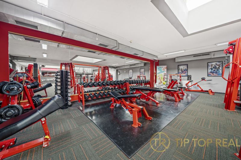 Fitness centrum s posilovacími zařízeními, lavicemi a zrcadly v obchodních prostorách.