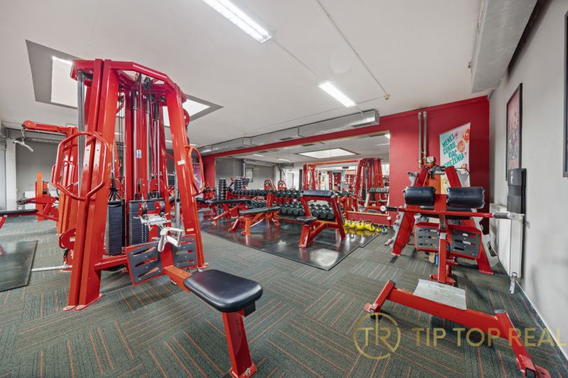 Fitness centrum s červenými posilovacími stroji a činkami, obchodní prostory.
